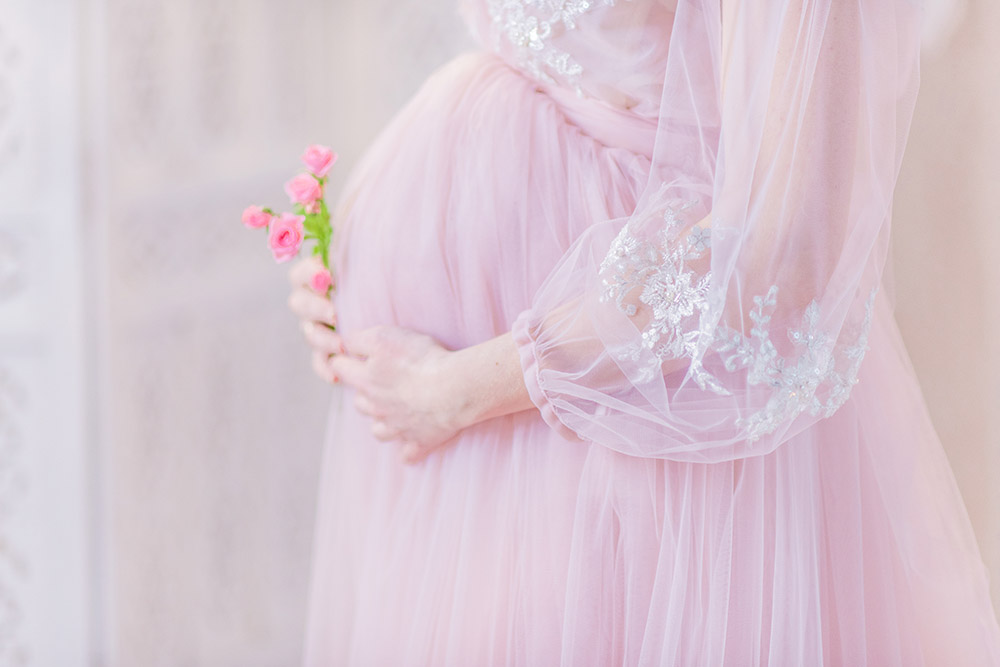 Beautiful pregnant woman in rich pink dress holds hands on her belly posing in luxury white room беременность без проблем